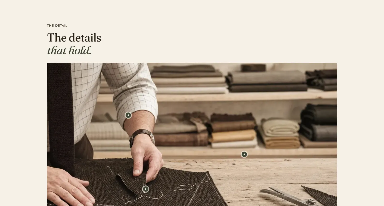 The Lapels Image hotspot section: the label "The detail" over the heading "The details that hold." with the last two words in italic, above a photograph of a tailor's hands pinning dark herringbone cloth on a workbench in front of stacked bolts of cloth, with three small round marks set on the cloth, the shears and the folded stack.