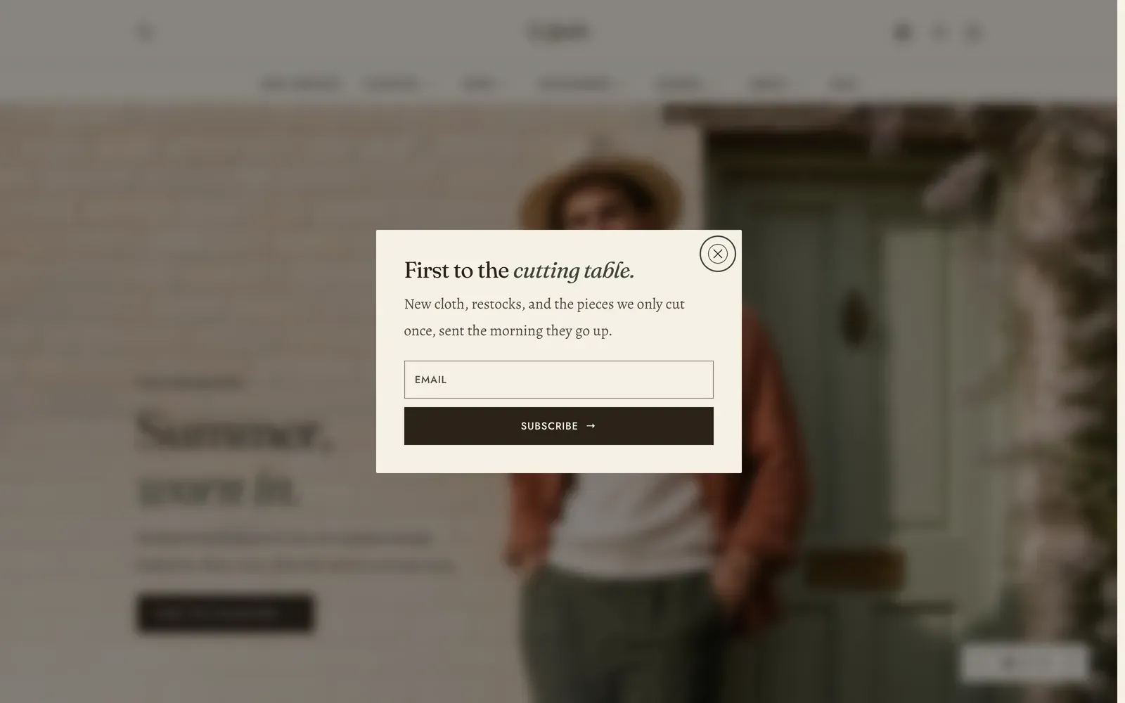 The Lapels newsletter popup in its centered layout, open over a dimmed and blurred home page: a small pale panel carrying the heading First to the cutting table with its second half in italic, a line about new cloth and restocks, an email field and a dark Subscribe button, with a round close control at its top right.