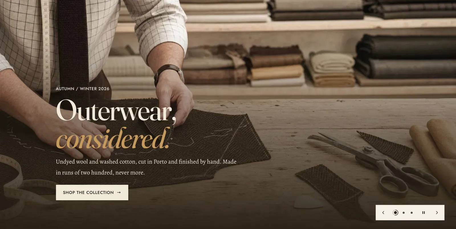 The Lapels Slideshow section: a full-bleed photograph of a cutter chalking a pattern onto dark cloth beside shears on a workbench, washed dark from the bottom, carrying the label "Autumn / Winter 2026", the heading "Outerwear, considered." with the last word in italic, a line about undyed wool cut in Porto, and a "Shop the collection" button. In the lower right a pale control bar holds a back arrow, three dots whose first carries a progress ring, a pause control and a forward arrow.